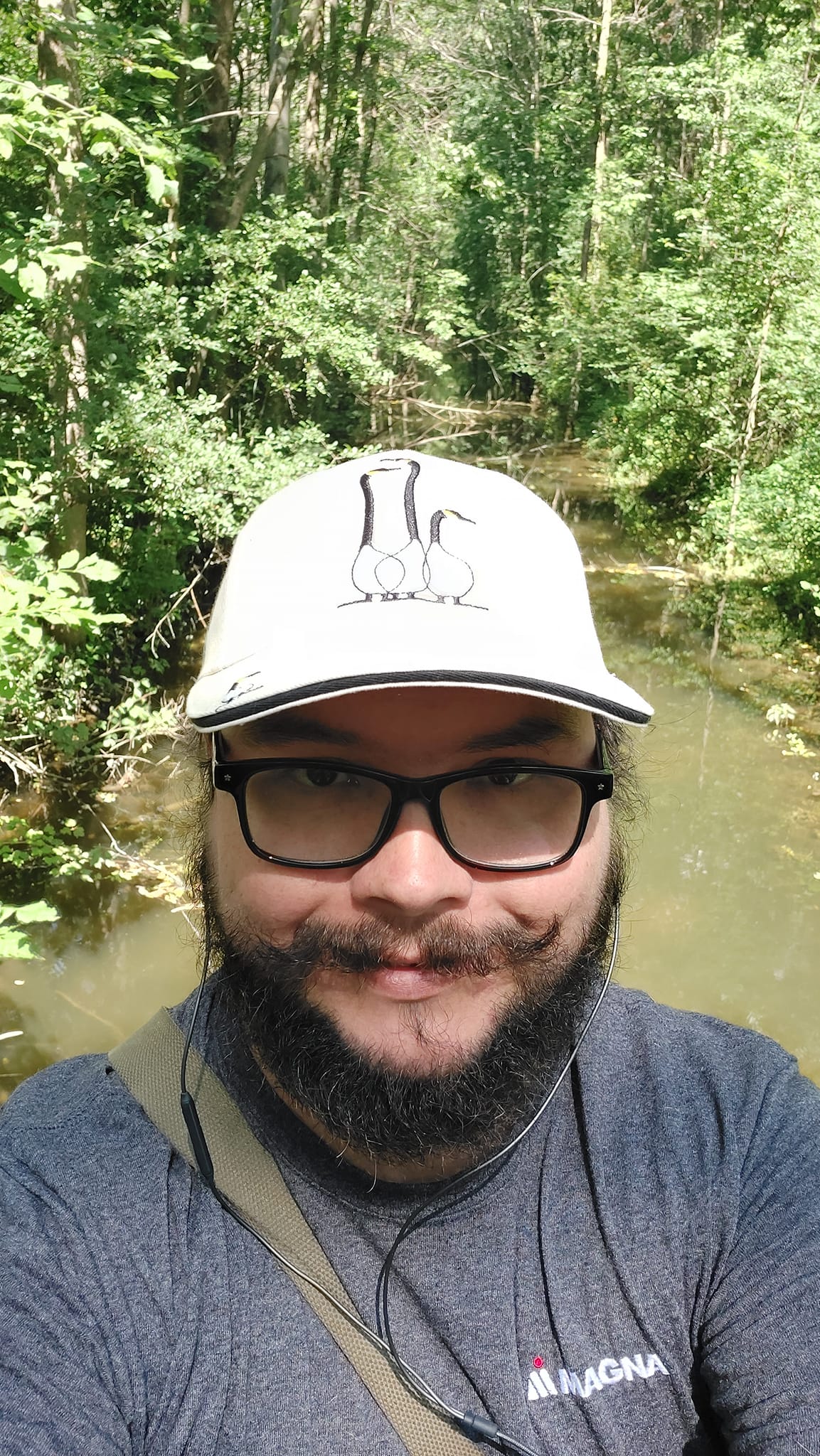 A man with short hair and bushy facial hair stands in front of a drainage ditch on a dam, the water is murkey